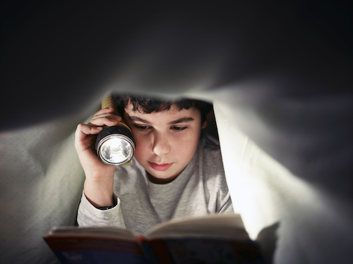 a boy reading a book under covers at night